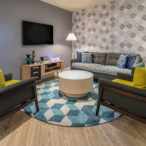 A modern living room features a sofa, two armchairs with yellow pillows, a round coffee table, a wall-mounted TV, and geometric wall décor.