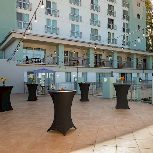 This image shows an outdoor patio area with high-top tables covered in black cloth, decorated with flowers in vases. A multi-story building is visible.