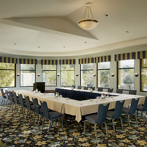 A conference room with a U-shaped arrangement of tables and chairs, surrounded by large windows and detailed carpet, is empty and well-lit.