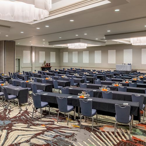 The image depicts a large conference room set up with rows of tables and chairs, ready for attendees. The room has a modern design with carpet flooring.