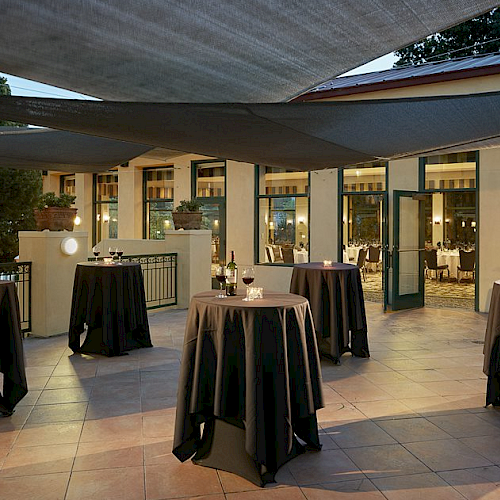 An outdoor event setup with cocktail tables covered in black tablecloths, lit candles, and a shaded canopy overhead on a patio.
