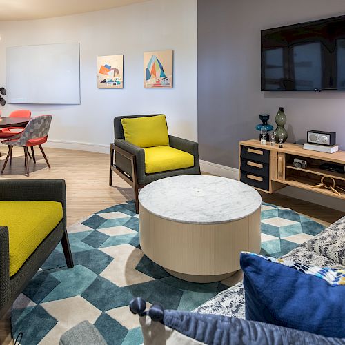 A modern living room with yellow-accented chairs, a round coffee table, a wall-mounted TV, and a dining area with a clock and colorful artwork.