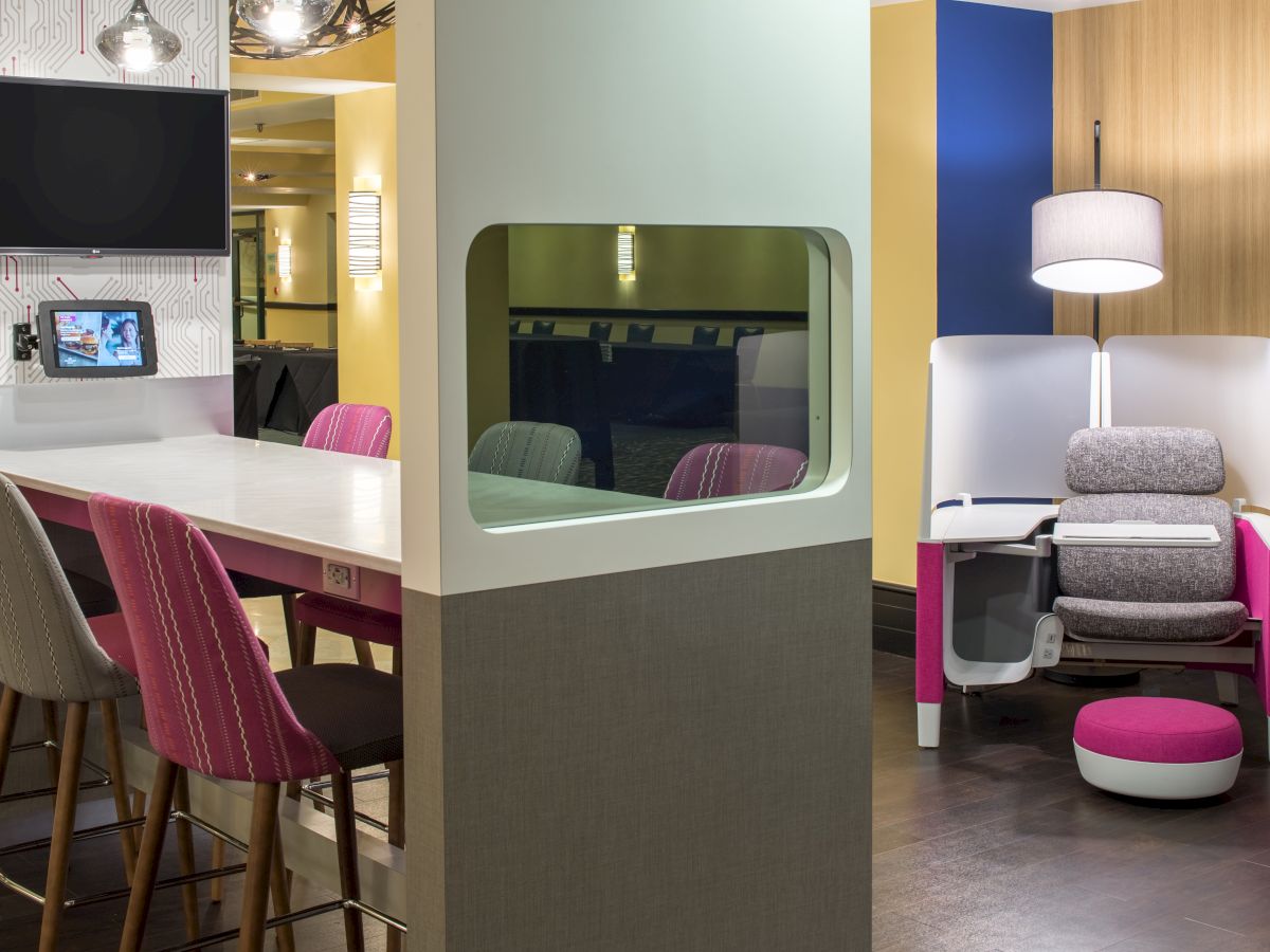 A cozy workspace with modern seating, a high table with pink chairs, a partitioned seating area, stylish lighting, and wall-mounted screens.
