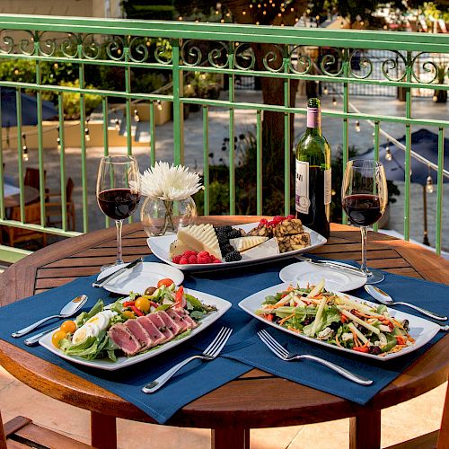 A balcony table set for two with salads, red wine, and assorted cheeses and fruits, with a scenic outdoor view.