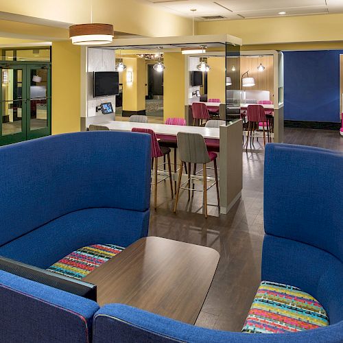 The image shows a brightly lit lounge area with blue curved seating, high tables with chairs, colorful decor, and modern lighting fixtures.