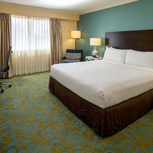 A hotel room with a king-sized bed, nightstands with lamps, a TV, a window with curtains, and a desk and chair. The carpet has a colorful pattern.