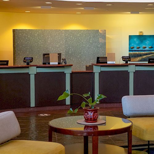 The image shows a hotel reception area with four counters, a seating area with chairs and a table with a plant on it, and artwork on the wall.