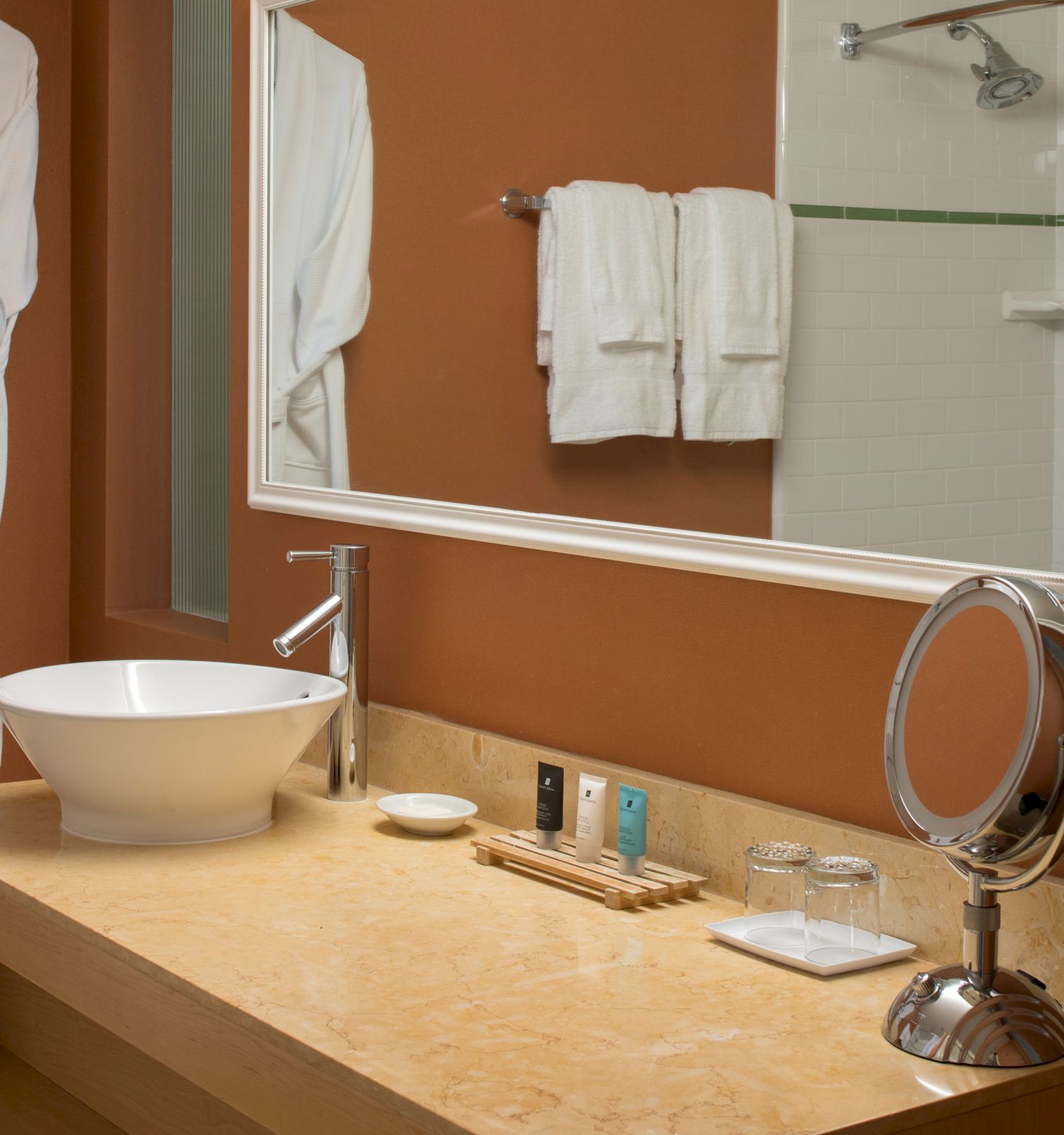 A bathroom with a countertop sink, a vanity mirror, toiletries, a bathrobe on a hook, two towels on a rack, and a shower visible in the background.