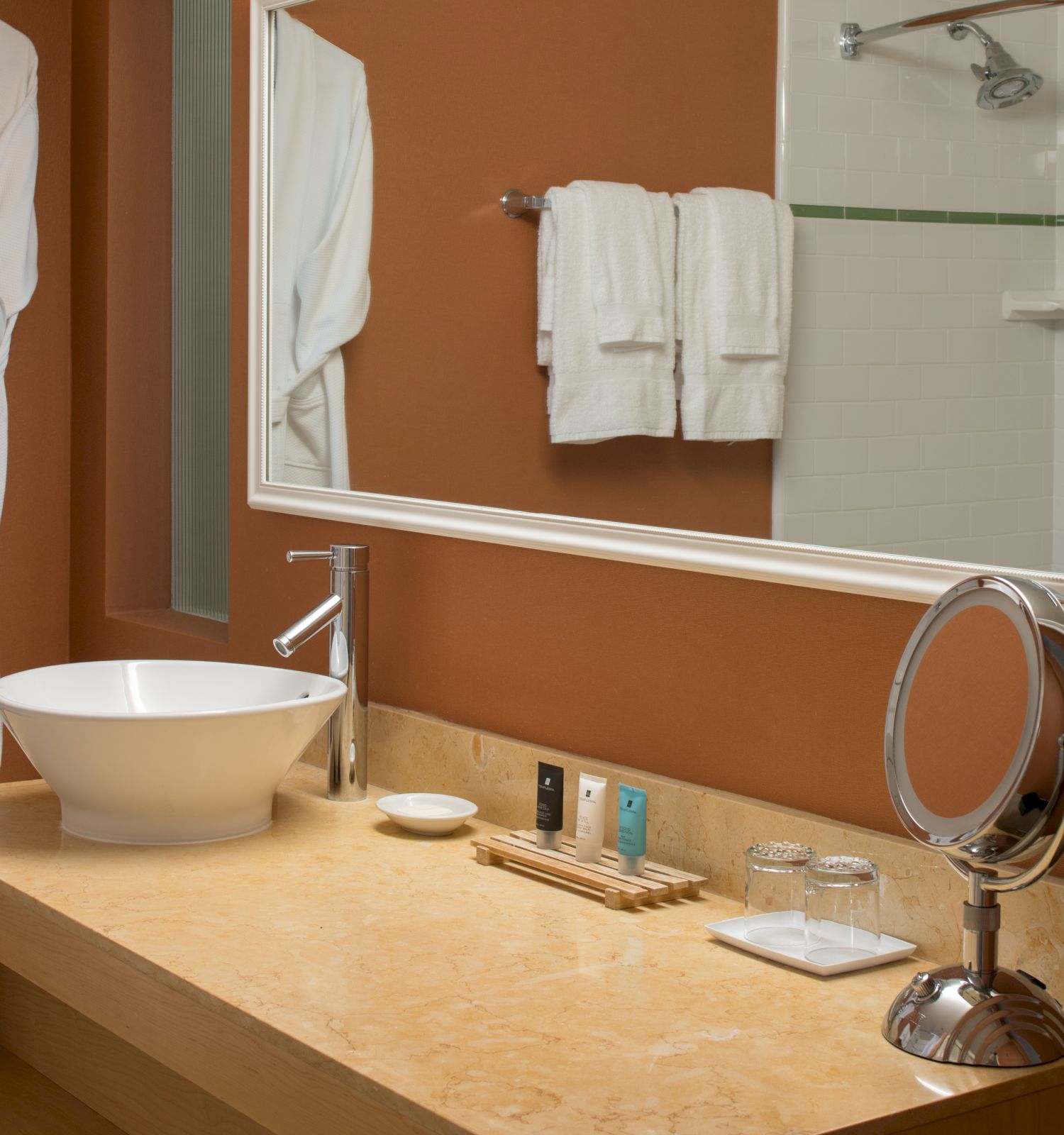 A bathroom with a countertop sink, faucet, mirror, round magnifying mirror, toiletries, towels, and a hanging bathrobe against a tan wall.