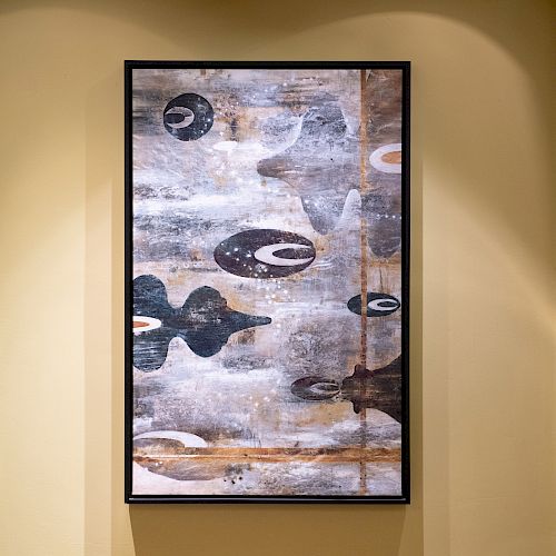 The image shows a framed abstract painting with a mix of earthy tones, shapes, and textures hung on a beige wall.