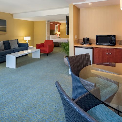 A modern hotel room with a sitting area, sofa, red chair, kitchenette, and a dining table with chairs. The space appears clean and well-lit.