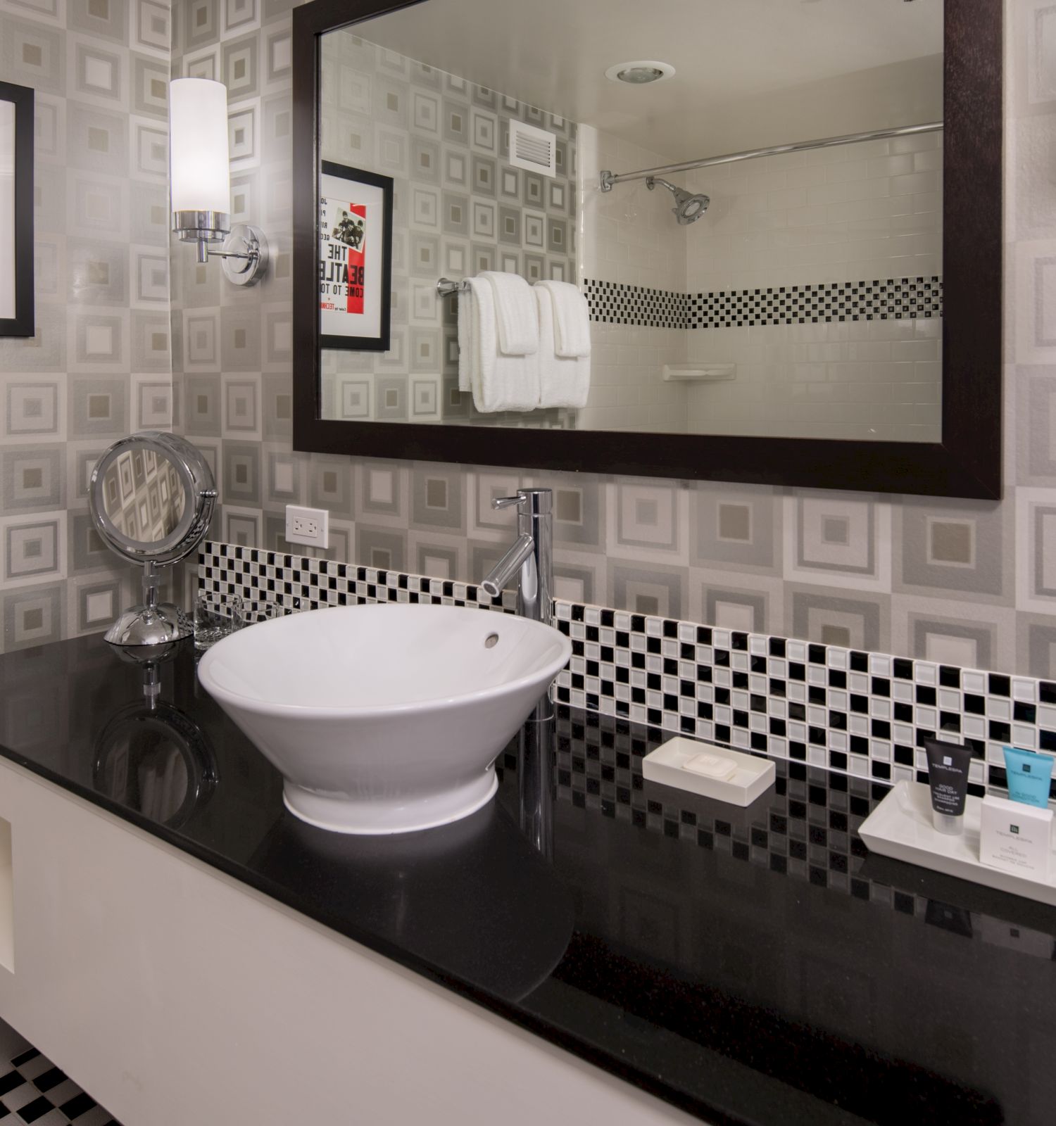 A modern bathroom features a white sink on a black countertop, a large mirror, a wall art piece, and geometric patterned tiles.