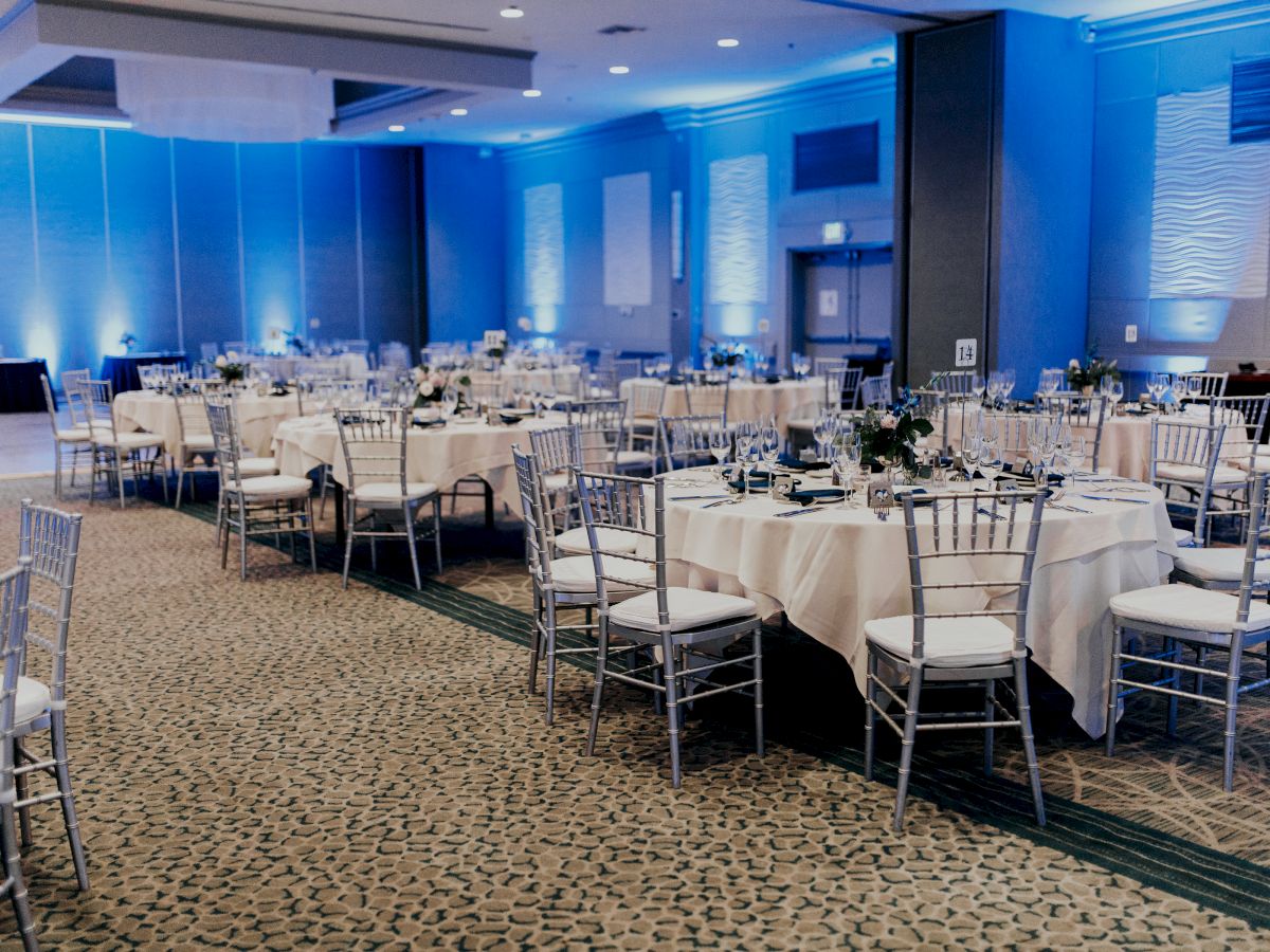 The image shows a banquet hall with round tables set up for an event, decorated with white tablecloths and chairs, and blue ambient lighting.