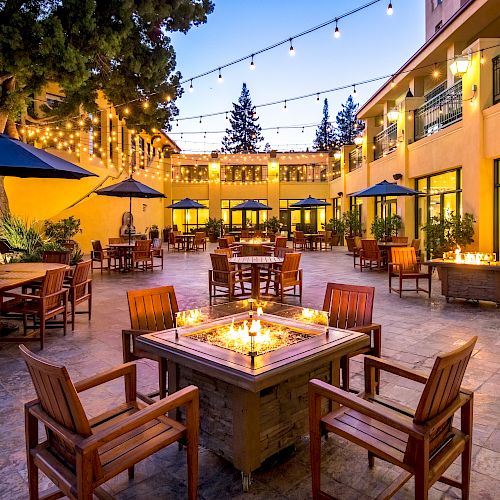 An outdoor courtyard with wooden tables and chairs, fire pits, string lights overhead, and yellow-lit buildings. Trees and umbrellas enhance the ambiance.