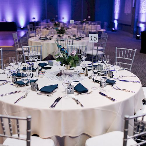 A decorated banquet hall with round tables set with white tablecloths, blue napkins, glassware, cutlery, and floral centerpieces, numbered as table 13.
