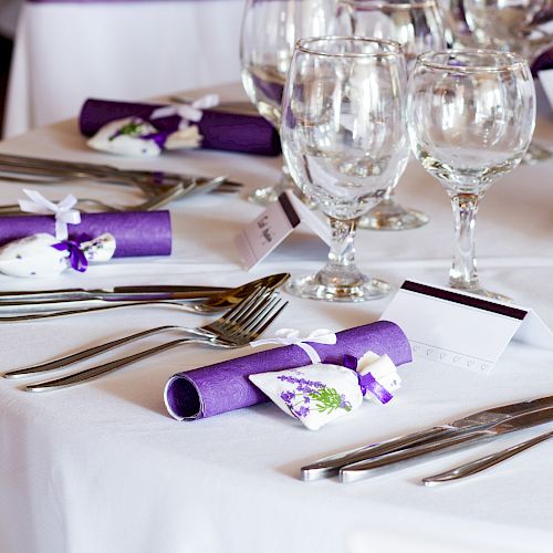 A elegantly set table with white tablecloth, purple napkins, glassware, and floral decor, featuring name cards and cutlery nicely arranged.