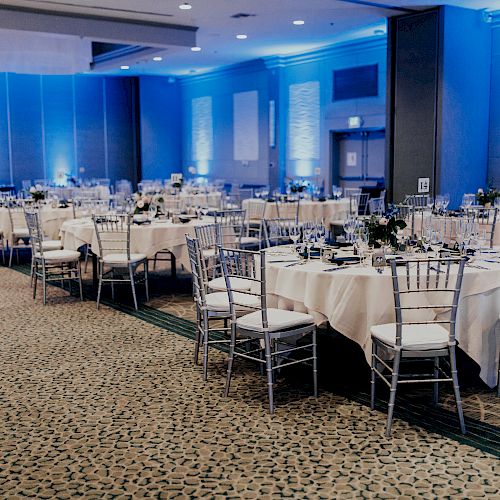 An elegantly decorated banquet hall with round tables covered in white tablecloths, silver chairs, and blue lighting, awaiting guests.