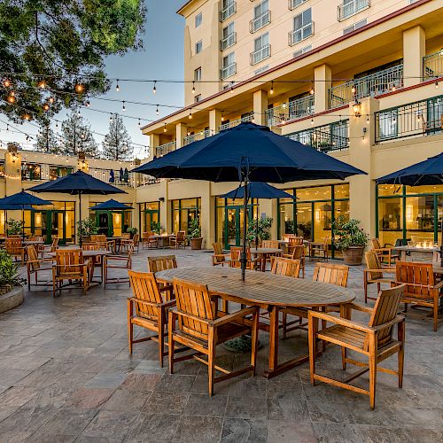An outdoor patio with wooden tables and chairs, blue umbrellas, string lights, and a multi-story building backdrop.