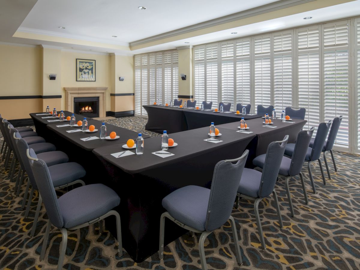 A conference room with a U-shaped table, chairs, notepads, pens, bottled water, and oranges, along with a lit fireplace and large windows with shutters.