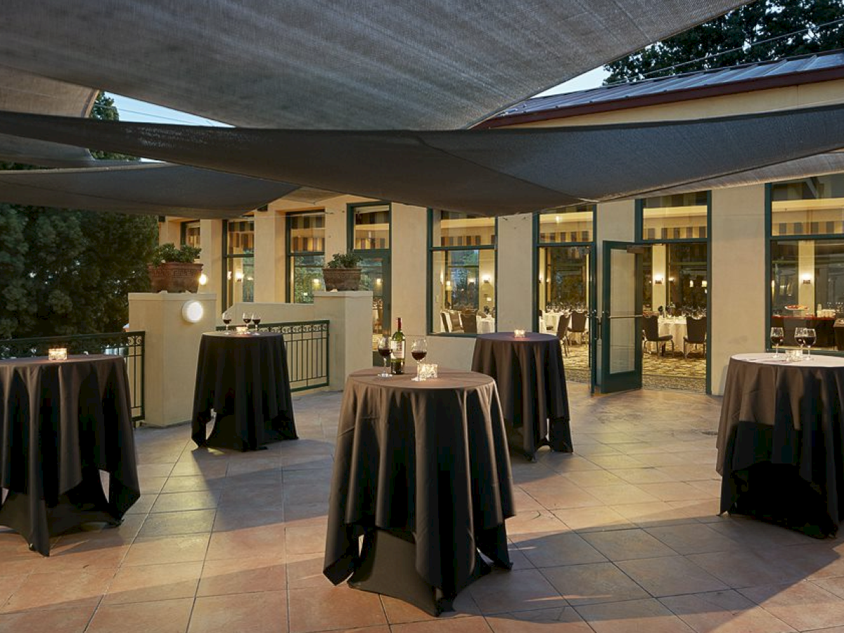 The image shows an outdoor event space with high tables covered in black tablecloths, candles, and some bottles, set under canopy shades.