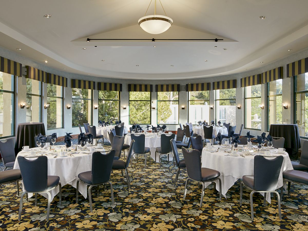 The image shows an elegant banquet hall with round tables set for dining, with white tablecloths, blue napkins, and chairs, surrounded by large windows.