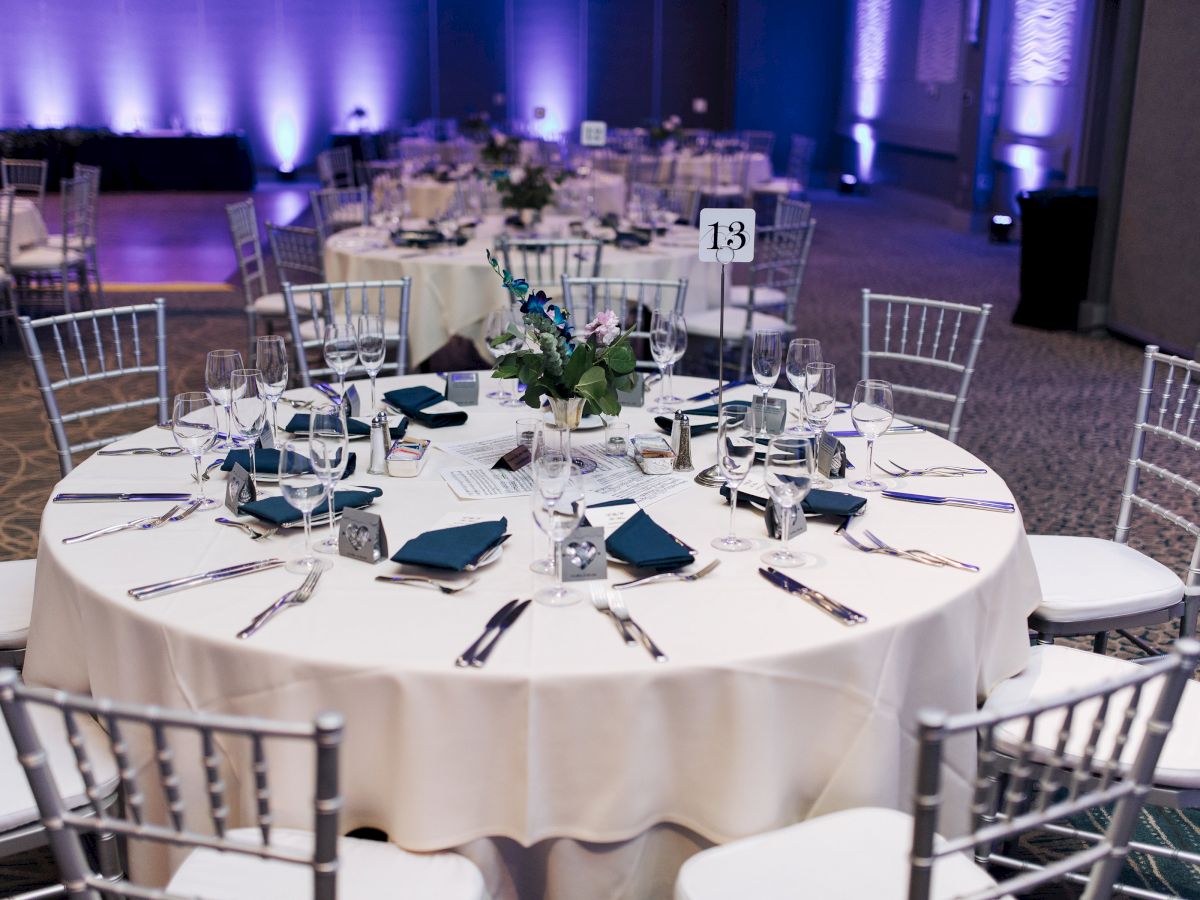 A round table set for an event, with elegant place settings, blue napkins, and a floral centerpiece, surrounded by silver chairs in a dimly lit hall.