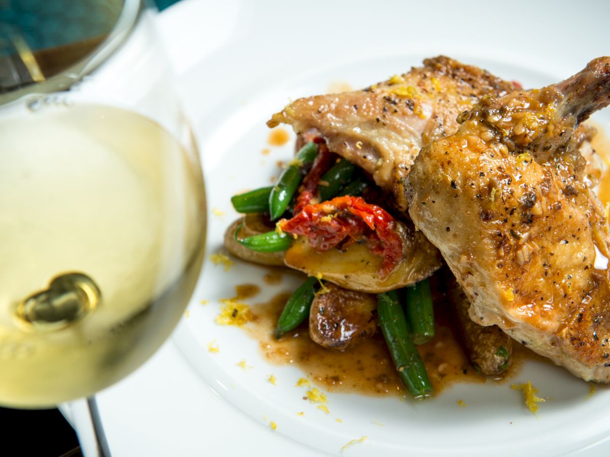 A plate of seasoned roasted chicken, green beans, and potatoes, served with a glass of white wine.