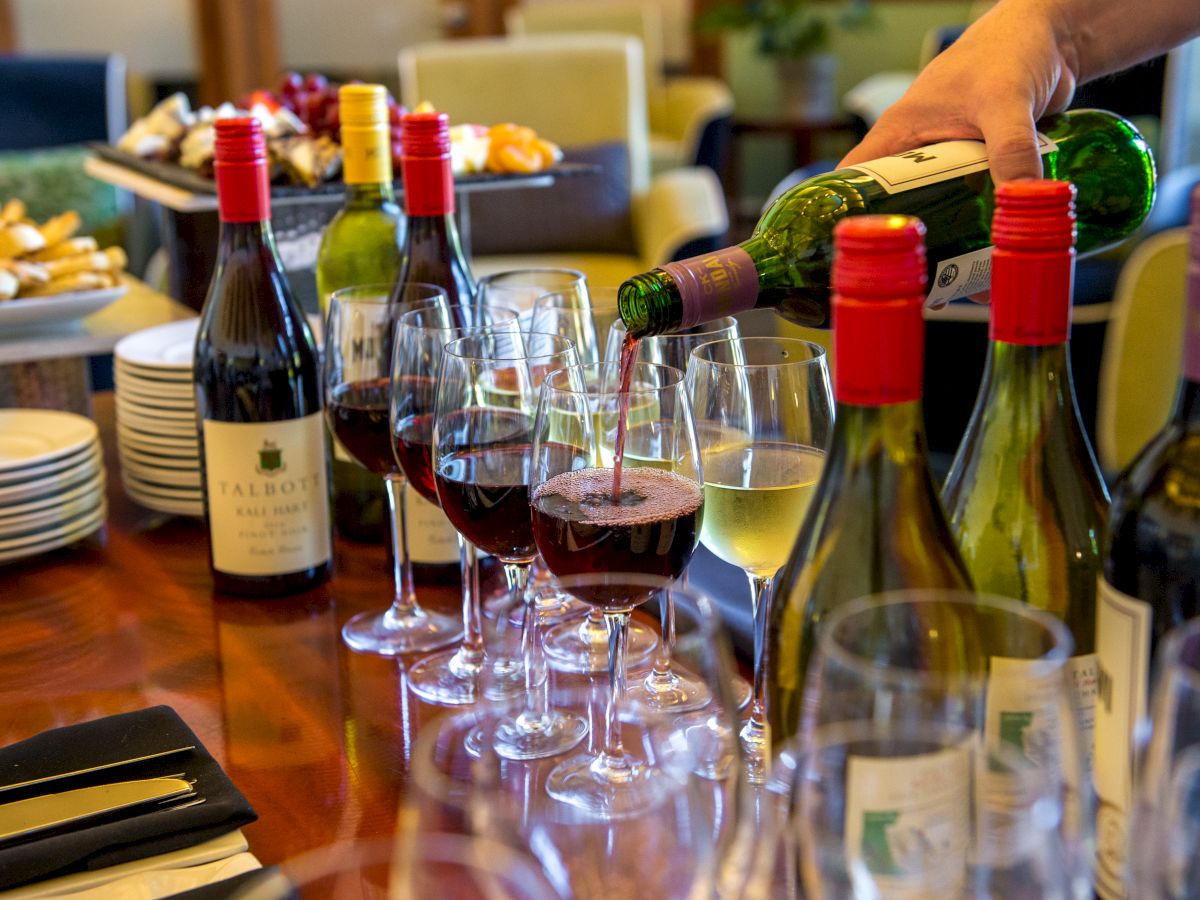 A person is pouring wine into glasses on a table set with various bottles of wine, wine glasses, and plates, likely prepared for a wine tasting event.