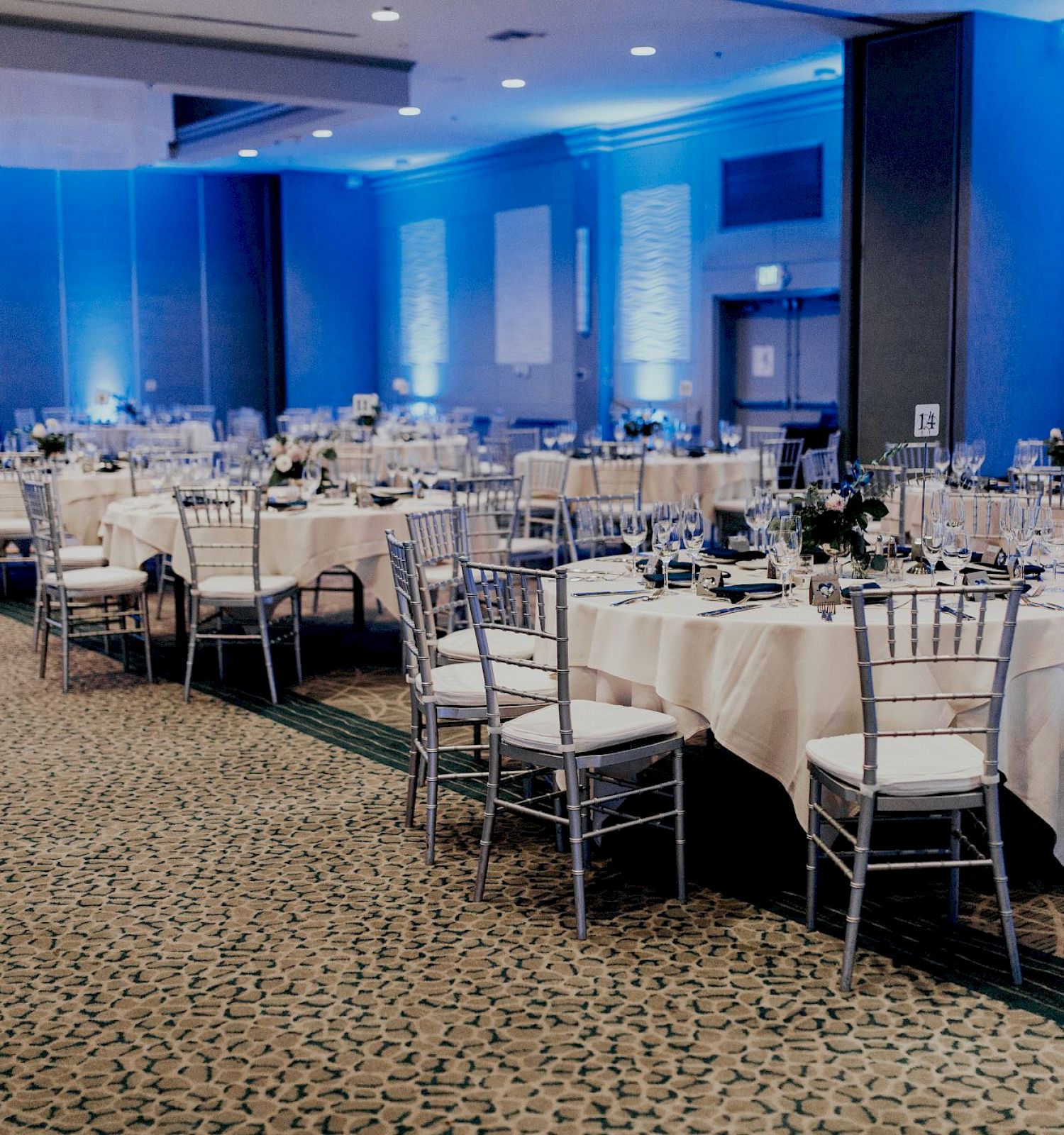The image shows an elegantly decorated banquet hall with round tables, white tablecloths, and silver chairs.