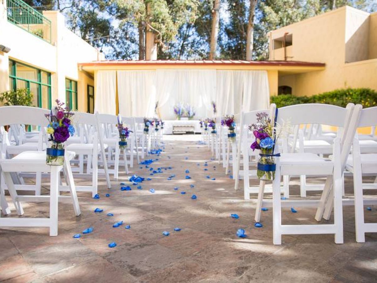 An outdoor wedding setup with white chairs, flower arrangements, and a white backdrop. Blue petals are scattered along the aisle, leading to the altar.