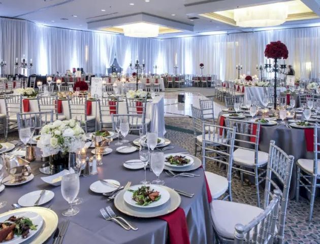 A well-decorated banquet hall with multiple round tables, silver chairs, place settings, floral centerpieces, and draped ceilings, ready for a formal event.