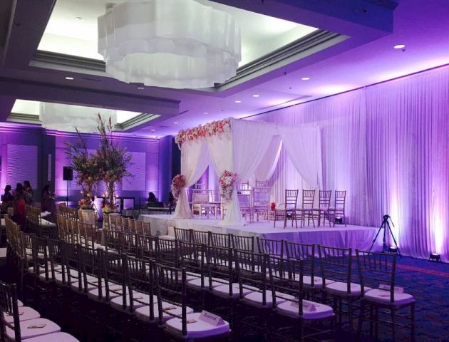 This image shows a decorated indoor wedding venue with rows of chairs, a flower arrangement, and a beautifully set stage with purple lighting.