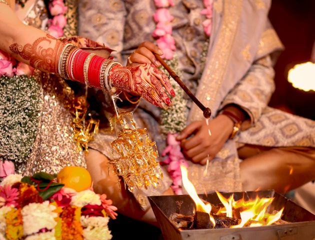 A wedding ceremony where a couple is performing rituals near a fire, with the bride's henna-decorated hands and jewelry visible.