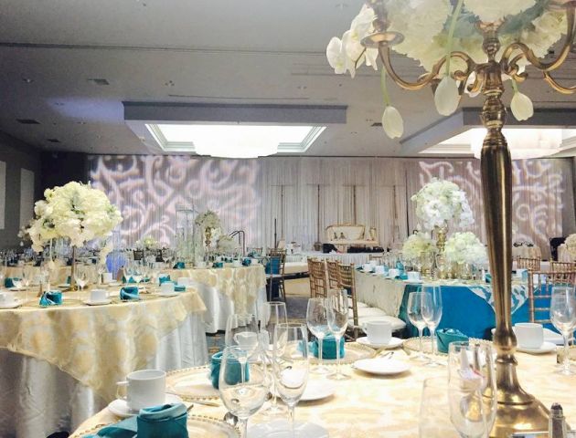 The image shows an elegant banquet hall with tables set up for an event, decorated with white flowers, blue napkins, and gold accents, under soft lighting.
