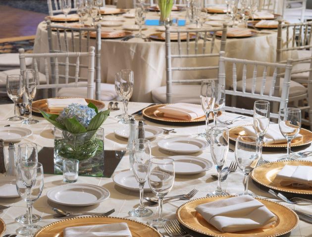 Elegantly set tables with white cloths and gold-rimmed plates, ready for a formal event, with floral centerpieces and neatly arranged cutlery.