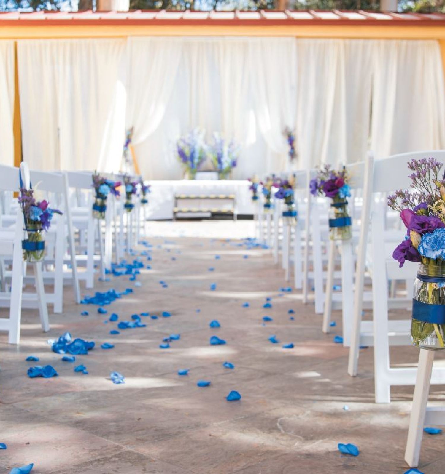 The image shows an outdoor wedding setup with white chairs adorned with floral arrangements and blue petals scattered on the aisle.