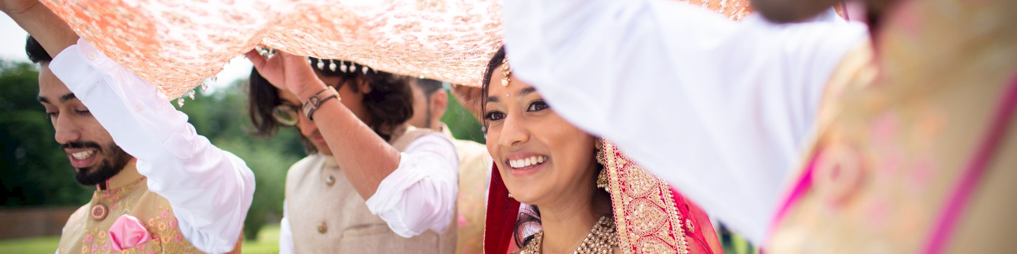 A bride walks under a decorated canopy, smiling, accompanied by three people in traditional attire, during an outdoor celebration or ceremony.