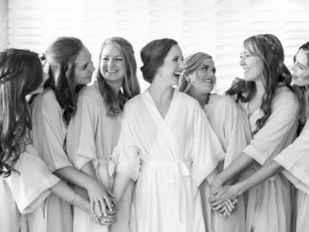 A group of women in robes stands close together, smiling and holding hands, appearing to enjoy a celebratory or intimate moment.