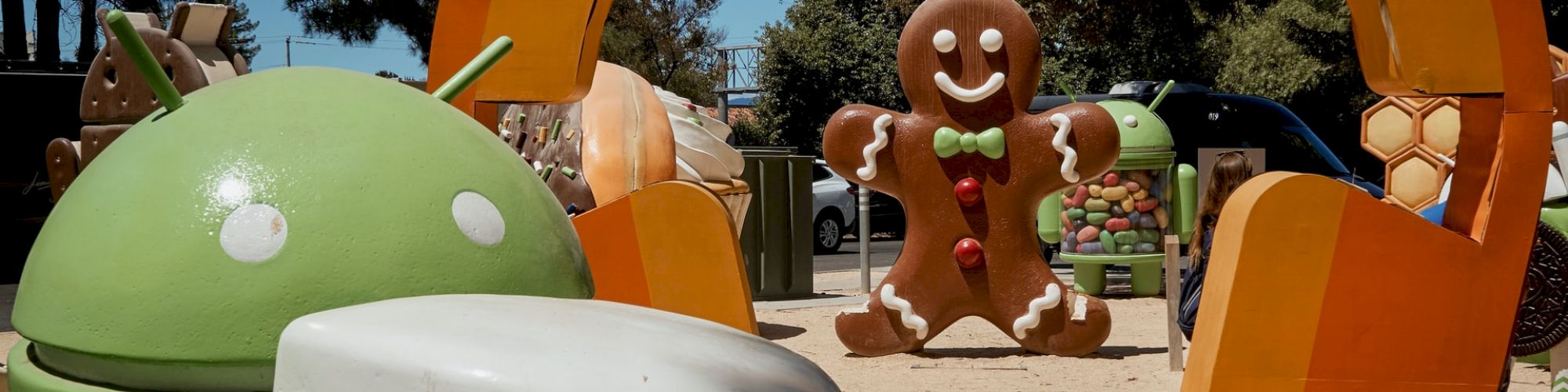 The image features Android mascot statues, including one holding a marshmallow and a large gingerbread figure, in an outdoor setting with trees in the background.