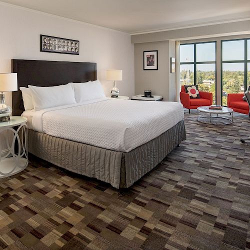 A modern hotel room features a king-size bed, two red chairs by large windows, a wall-mounted TV, and a turntable on a small table.