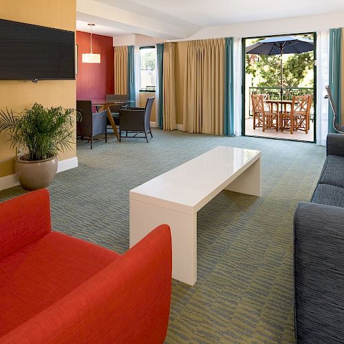 A spacious living room with a red chair, blue sofa, white coffee table, wall-mounted TV, dining area, and glass doors leading to an outdoor patio.