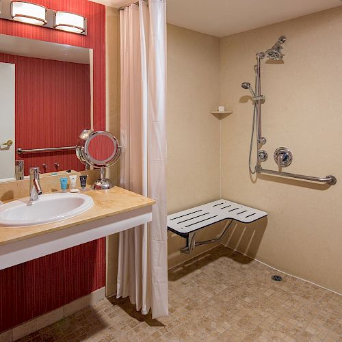 A clean and accessible bathroom with a sink, mirror, red walls, and a shower area with a seat and grab bars, designed for convenience.