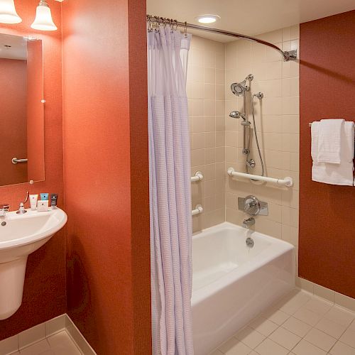 A bathroom with a sink, mirror, and toiletries on the left, and a bathtub with a shower curtain and towels on the right wall, always ending the sentence.