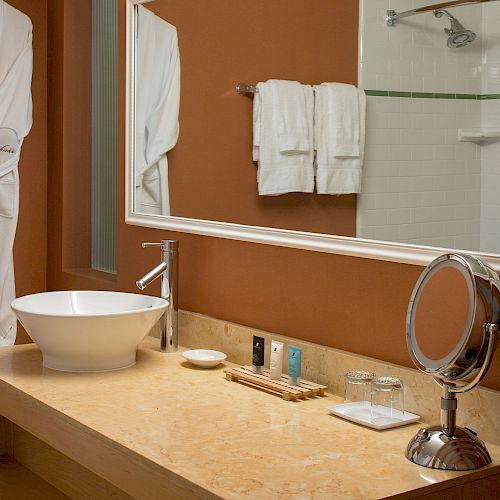 A bathroom with a robe hanging on the wall, a countertop with a vessel sink, toiletries, towels, a magnifying mirror, and a shower area in the background.