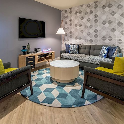 A modern living room with a patterned wall, a grey sofa, two armchairs with yellow cushions, a round coffee table, and a rug with geometric patterns.