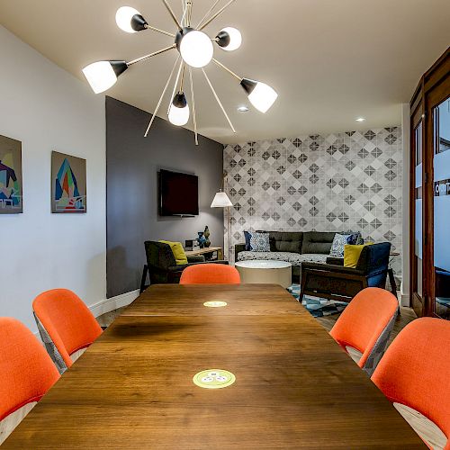 A modern conference room with a wooden table, six orange chairs, wall art, a television, and a cozy seating area in the background.