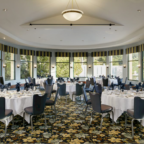 This image shows an elegantly set dining room with round tables, arranged chairs, and large windows, ready for an event or banquet.