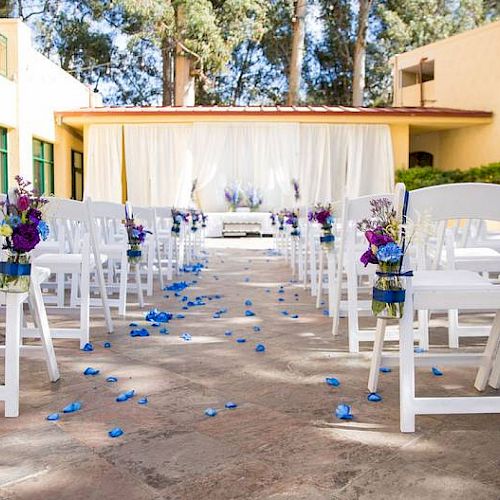 A wedding venue with white chairs and blue decorations, featuring flower arrangements and a white draped backdrop set outdoors on a sunny day.