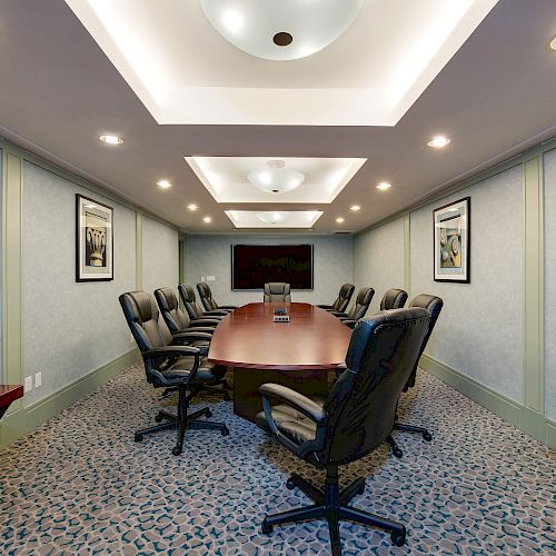 A modern conference room with a long wooden table, black office chairs, overhead lighting, light-colored walls, framed art, and a patterned carpet.