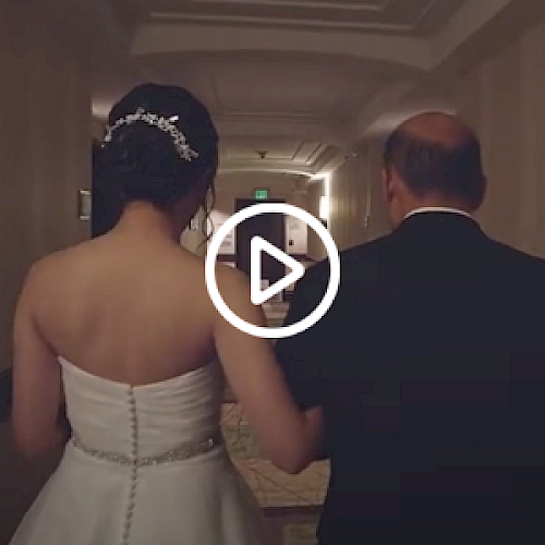 A bride and an older man walk down a hallway, with the video play symbol over them, suggesting a wedding moment or ceremony.